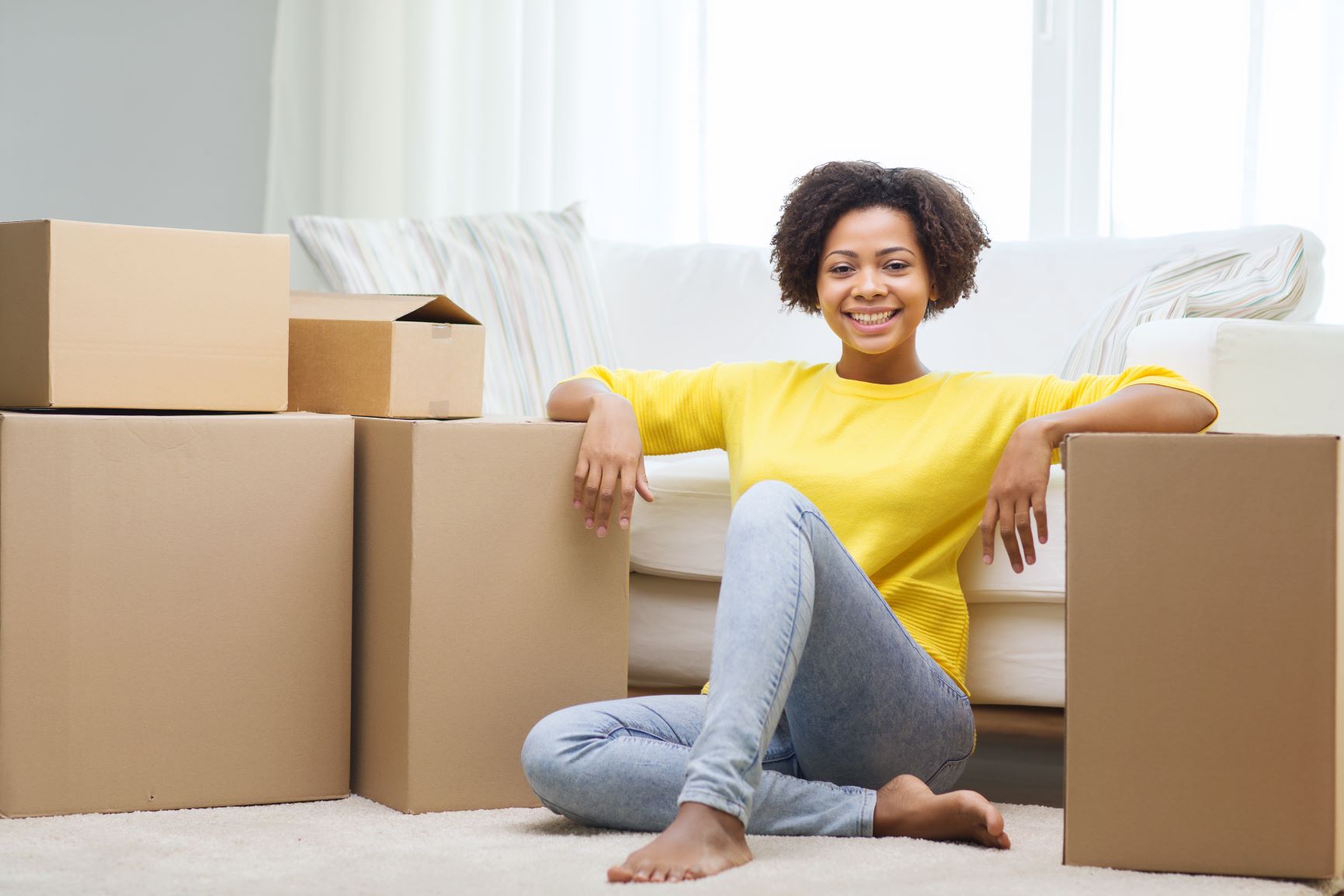 Woman Sitting by a couch and smiling while leaning against packed moving boxes
