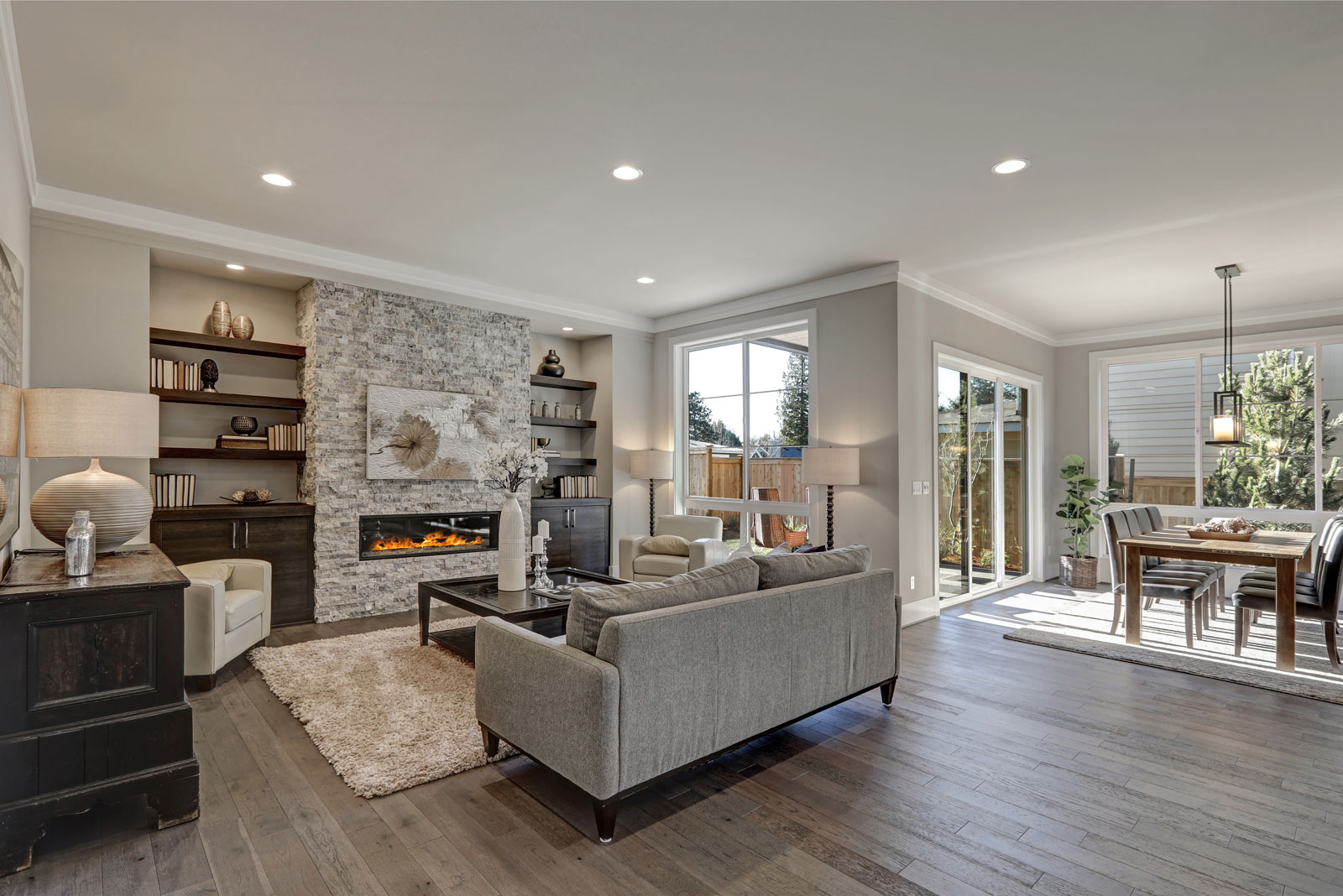 Living room interior in gray and brown colors features gray sofa atop dark hardwood floors facing stone fireplace with built-in shelves.